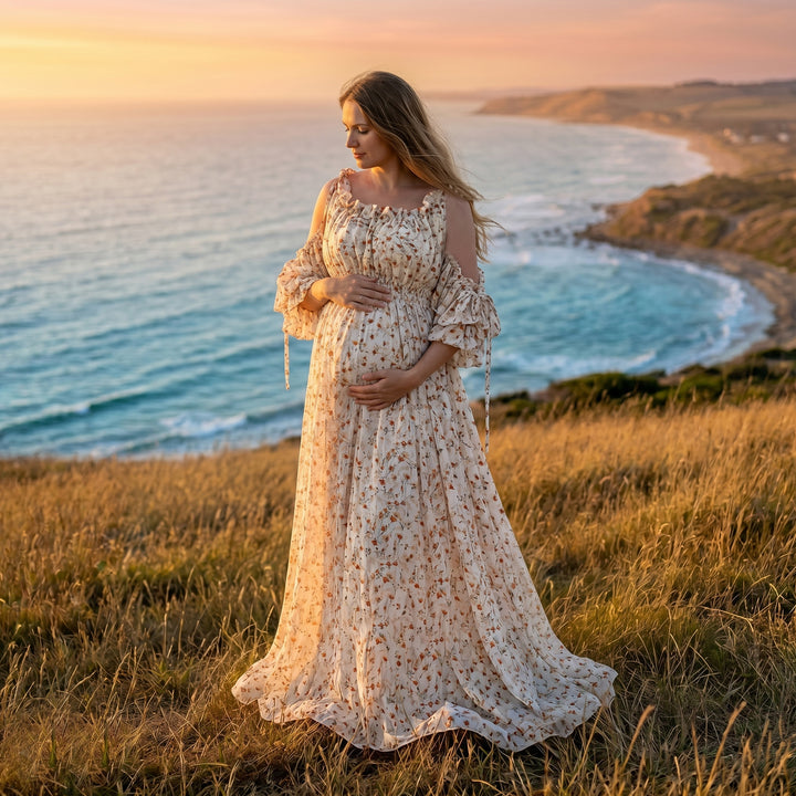 A pregnant woman in an Alke floral maxi dress stands on a grassy cliff by the sea, holding her belly.