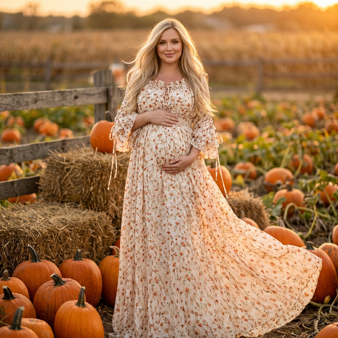 A pregnant woman in an Alke orange floral dress stands in a pumpkin patch holding her belly, with hay bales and a wooden fence nearby.
