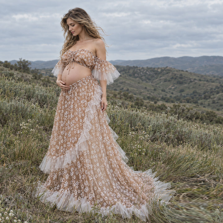 A pregnant woman stands outdoors in a grassy, hilly landscape wearing a sheer khaki dress with white floral embroidery, a matching off-the-shoulder crop top, and layered tulle ruffles.