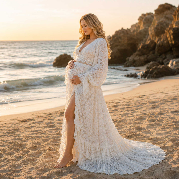 A pregnant woman wears the ivory lace gown standing on sand, with a side slit revealing her leg.