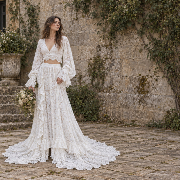 A woman stands outdoors in front of a stone wall and steps, wearing a ivory lace two-piece outfit with a long-sleeve cropped top and a full ruffled skirt with a train. 