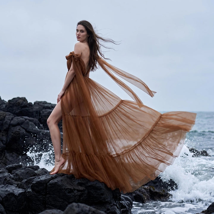 A woman in a Acaste gown stands on black rocks with the skirt blowing in the wind.