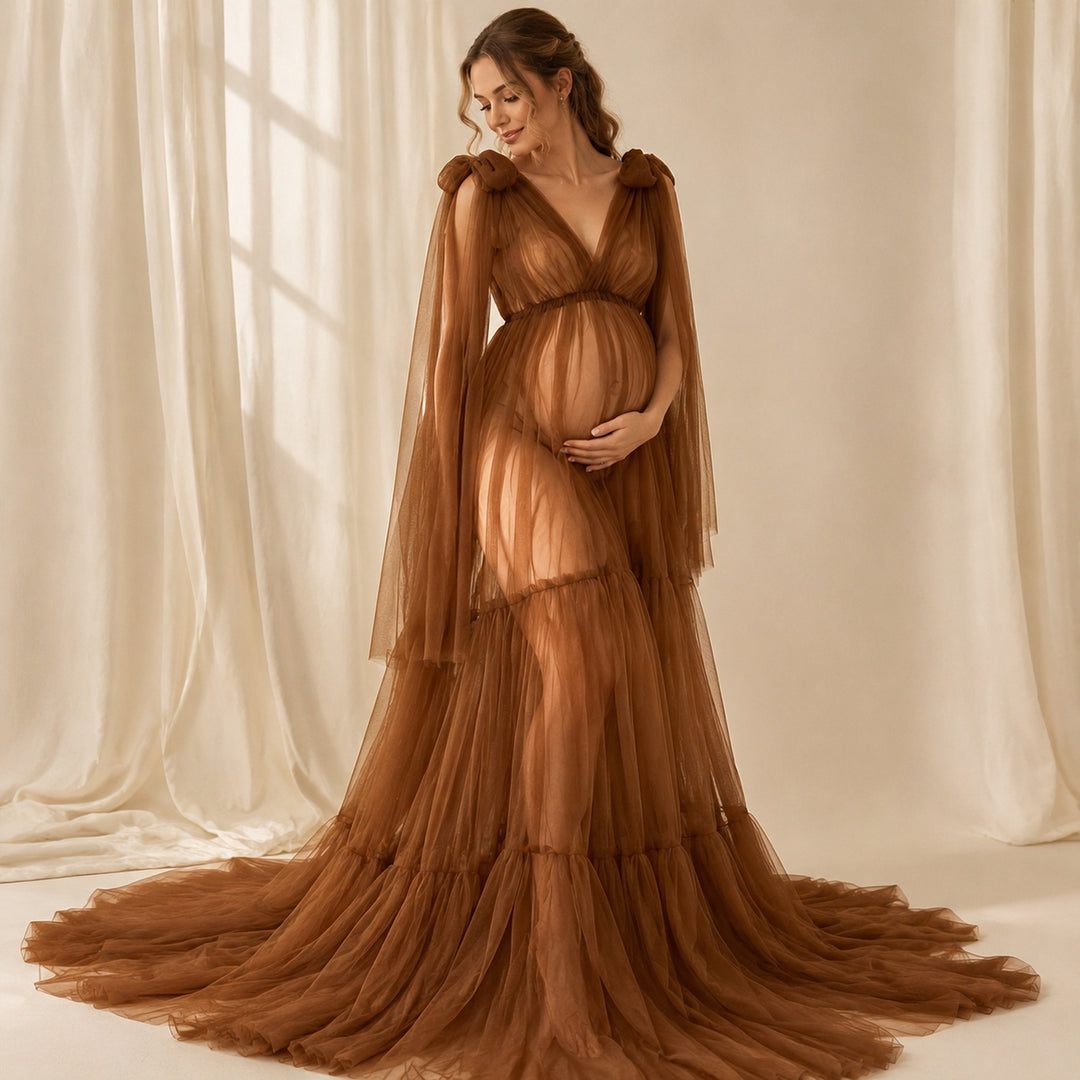 A pregnant woman in a Acaste gown, stands before white curtains, cradling her belly.