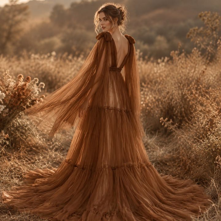 A woman in a Acaste gown, stands in dry grass, turning back to look over her shoulder; the train drags on the ground and blows in the wind.