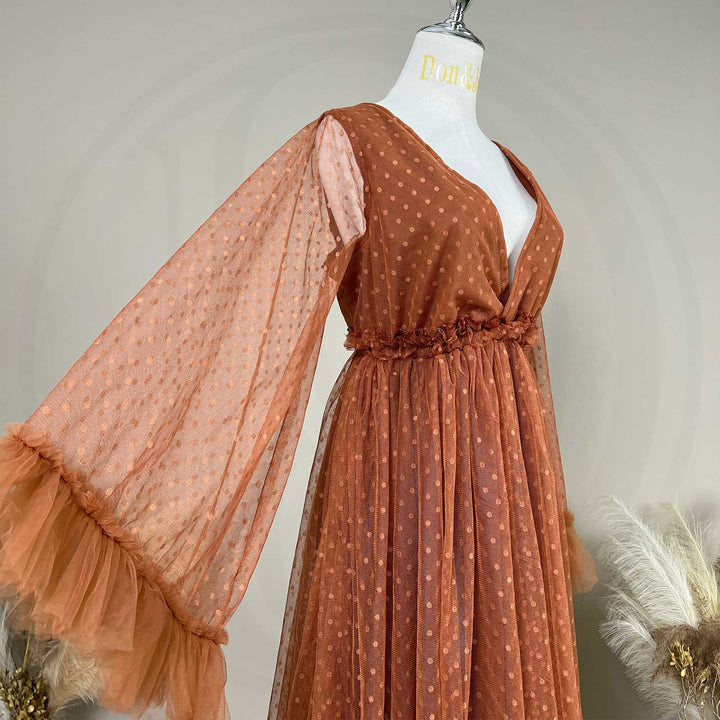 A terracotta polka dot tulle maternity dress is displayed on a mannequin torso. The dress features a V-neckline, long sheer sleeves with ruffled cuffs, and a gathered waist with ruffle detailing.