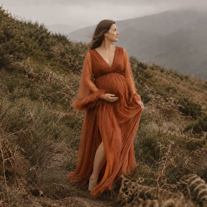 A pregnant woman stands on a misty, wearing a terracotta polka dot maternity gown with sheer long sleeves,ruffled cuffs, a V neckline.