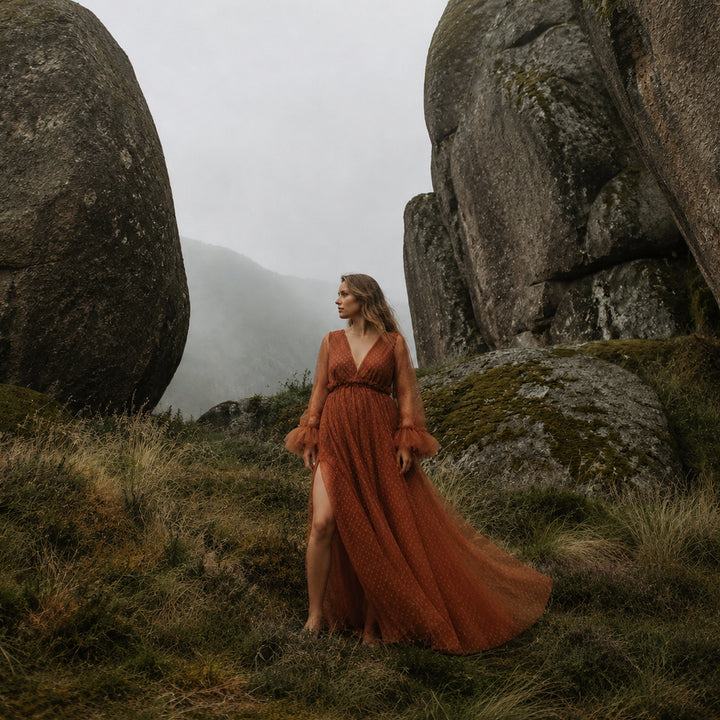 A pregnant woman stands between large boulders on a grassy hillside wearing a terracotta polka dot tulle maternity gown with long ruffled sleeves, a V-neckline, and a high side slit.