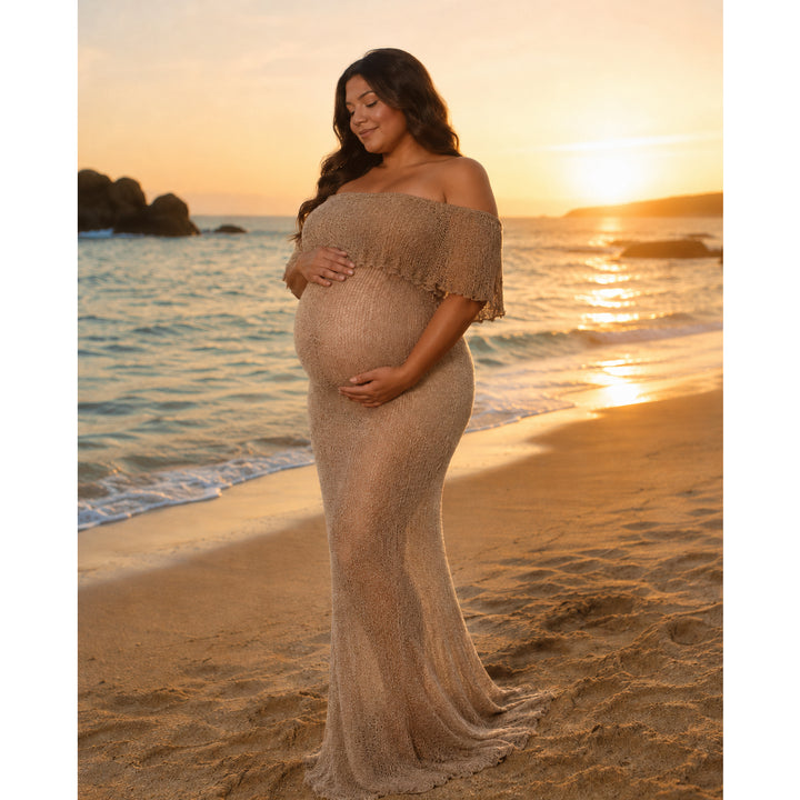 A pregnant woman in an Aletheia  camel sparkly gown stands on the beach, hands on her belly.