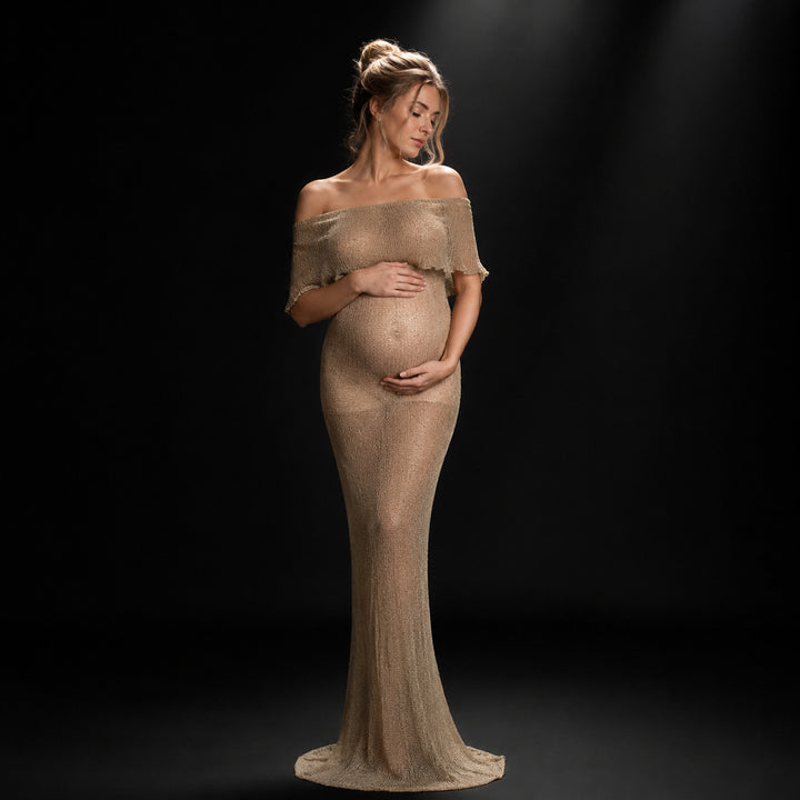 A pregnant woman in an Aletheia  camel sparkling off-shoulder gown stands holding her belly.