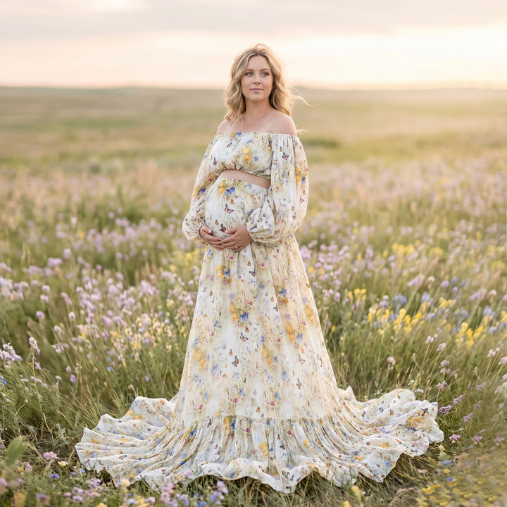 A pregnant woman in an Aphaea floral off-shoulder two-piece stands in a flower field holding her belly.