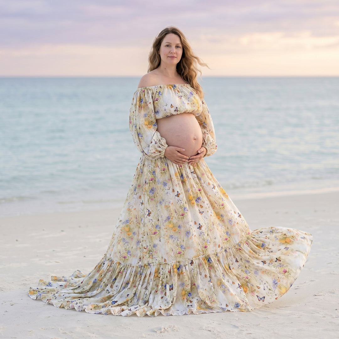 A pregnant woman in an Aphaea floral off-shoulder two-piece stands on the beach holding her belly.