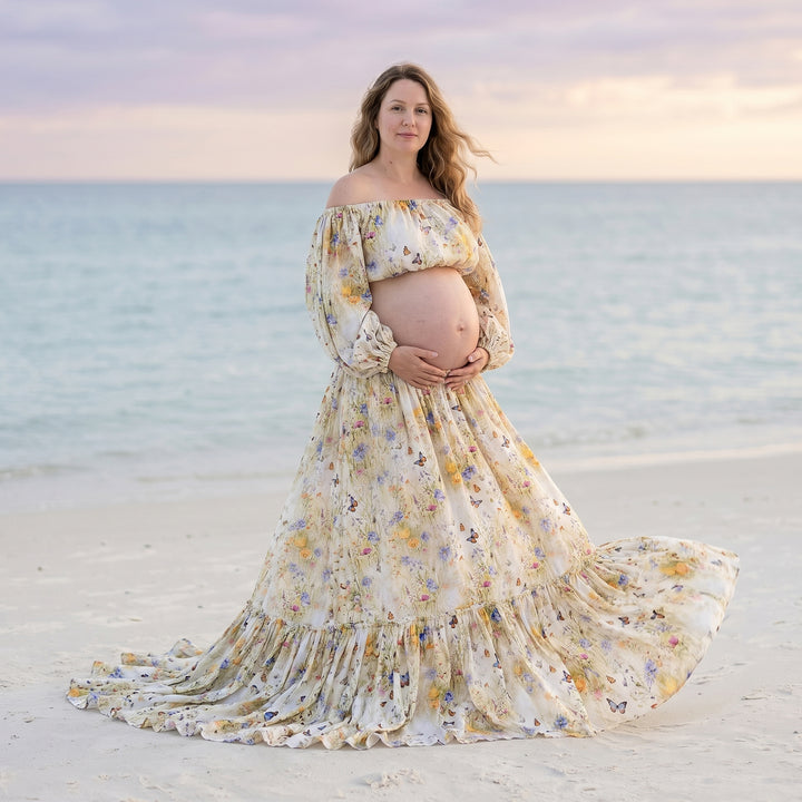 A pregnant woman in an Aphaea floral off-shoulder two-piece stands on the beach holding her belly.