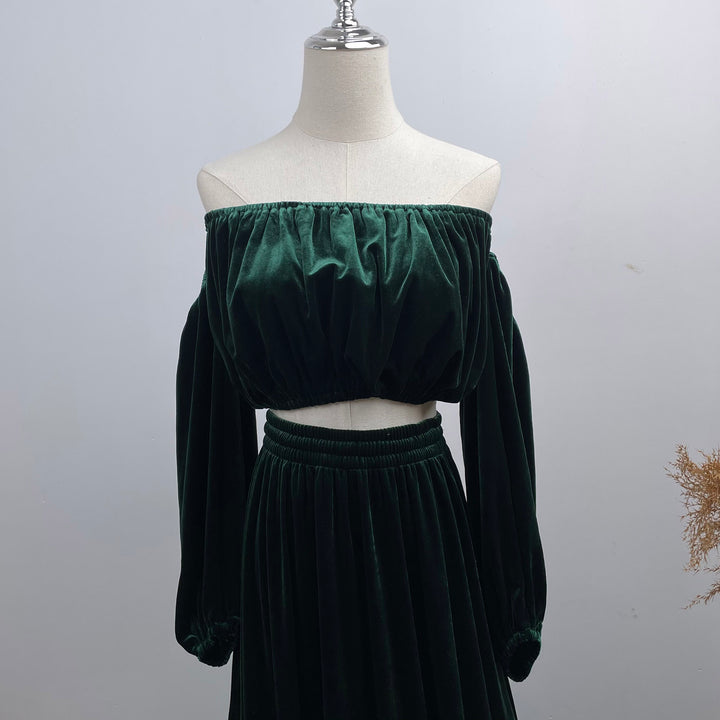 A dark green velvet off-the-shoulder two-piece set consisting of a cropped long-sleeve top and a matching high-waisted skirt, displayed on a cream dress form mannequin against a light gray background.