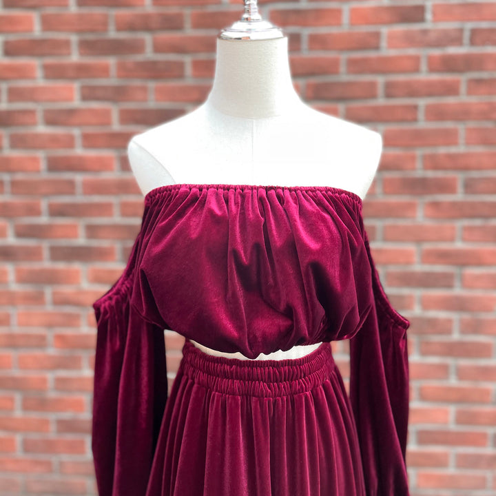 A deep burgundy velvet off-the-shoulder crop top and matching skirt set displayed on a white dress form mannequin, set against a red brick wall background.