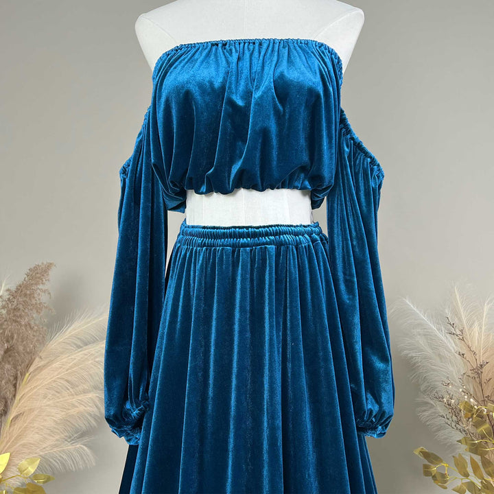A two-piece velvet two-tone dress in a rich blue color, displayed on a white mannequin. The set includes an off-the-shoulder long-sleeve crop top and a matching high-waisted maxi skirt.