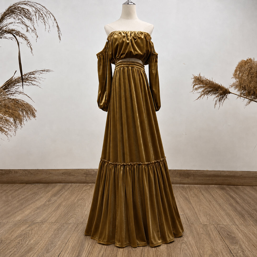 An off-the-shoulder long-sleeve maxi dress in a rich gold velvet fabric, displayed on a mannequin against a neutral wall with dried pampas grass accents.