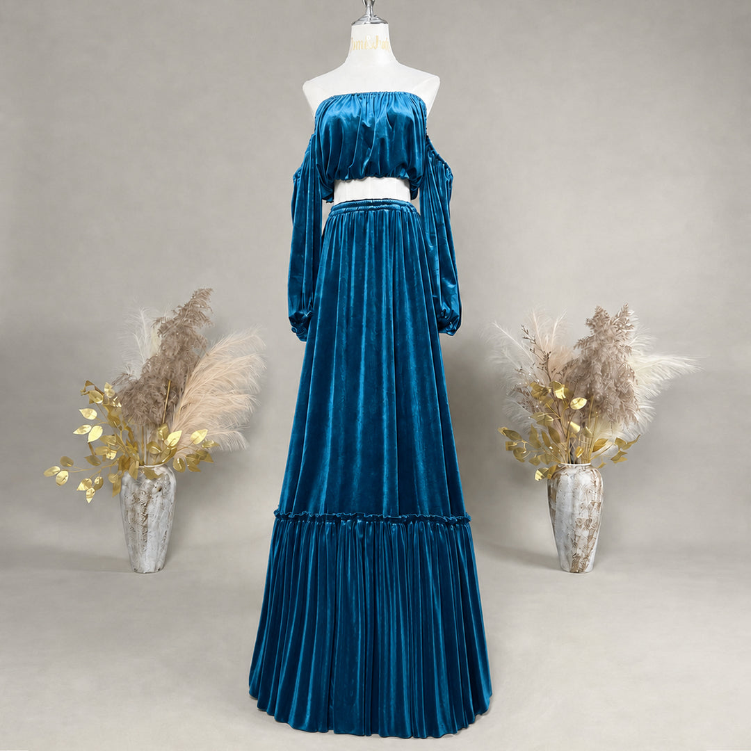 A long velvet off-the-shoulder maxi dress displayed on a mannequin against a neutral gray studio background, flanked by two decorative vases with dried grass and gold leaves.