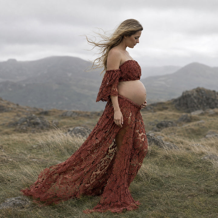 A pregnant woman stands in profile on a windswept grassy hill wearing a terracotta two-piece lace maternity set with off-the-shoulder ruffled sleeves and a high-slit skirt that reveals her bare baby bump against a mountain backdrop.