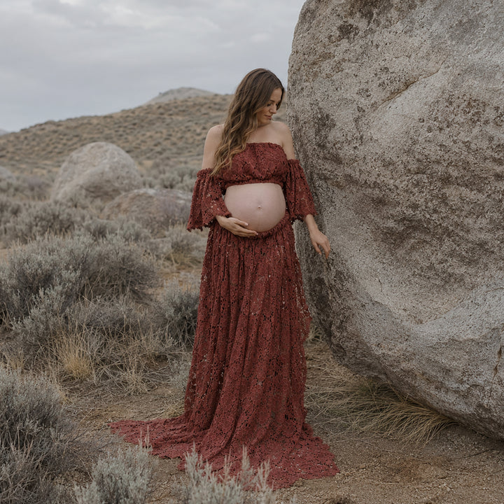 A pregnant woman stands beside a large boulder in a desert landscape wearing a terracotta off-the-shoulder lace maternity gown with ruffled sleeves and an open front skirt that reveals her baby bump.