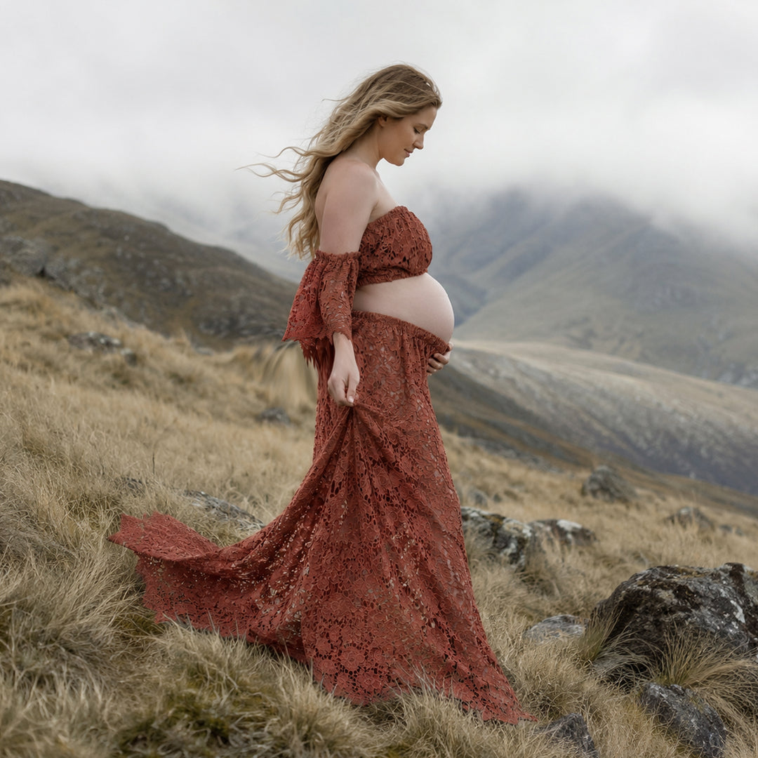 A pregnant woman stands in profile on a misty, grassy hillside wearing a terracotta two-piece lace maternity set with off-the-shoulder ruffled sleeves and a high-waisted skirt that reveals her bare baby bump against a mountain backdrop.