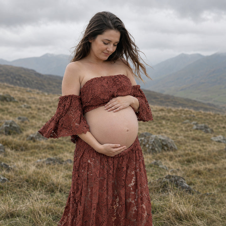 A pregnant woman stands in profile on a misty, grassy hillside wearing a terracotta two-piece lace maternity set with off-the-shoulder ruffled sleeves and a high-waisted skirt that reveals her bare baby bump against a mountain backdrop.