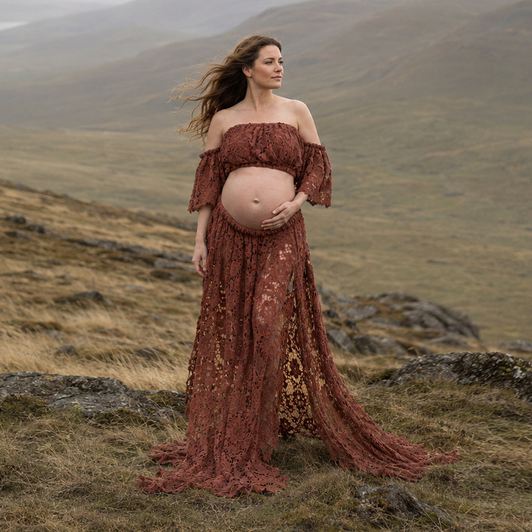 A pregnant woman stands on a grassy hillside wearing a Bendis terracotta off the shoulder lace maternity gown with ruffled sleeves and an open front skirt that reveals her baby bump and flows over the terrain.
