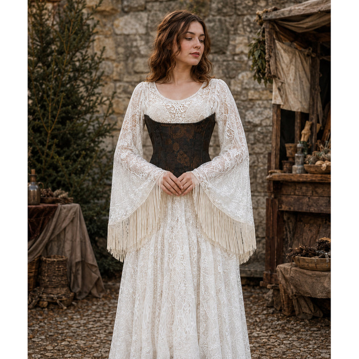 A woman stands outdoors in a rustic stone setting wearing a long white lace dress with a dark brown corset-style bodice and wide fringed sleeves.