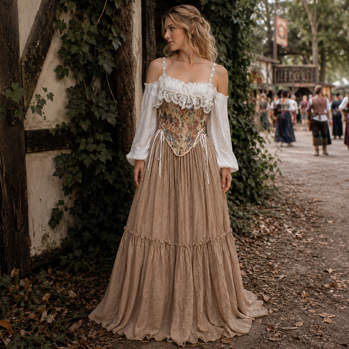 A woman stands outdoors by a stone wall wearing a Brigid ivory off-shoulder top, floral corset, and khaki skirt.