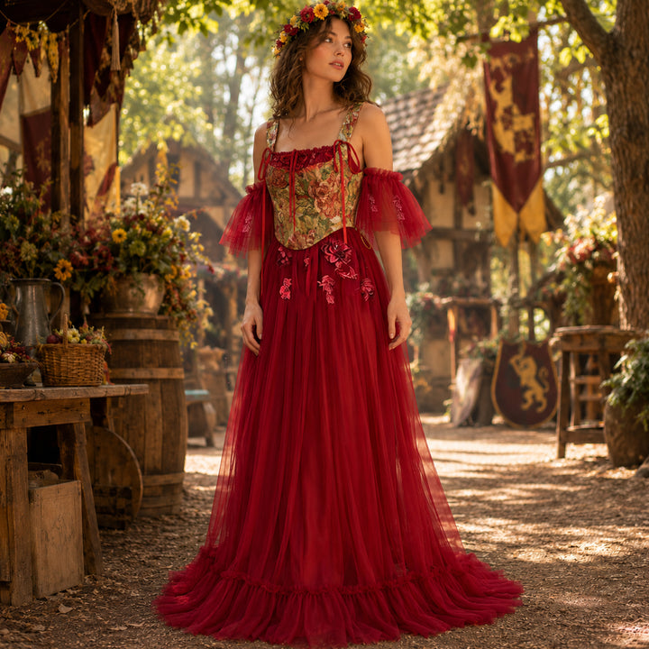 A woman wearing a Cailleach flower crown, red tulle gown, and floral corset stands in an outdoor medieval setting. Wooden houses, flags, and barrels with flowers are in the background.
