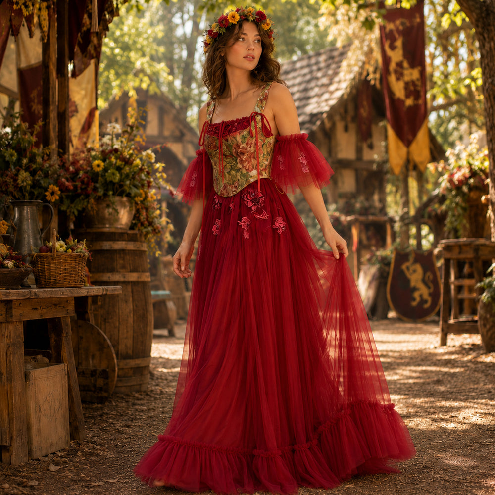 A woman wearing a Cailleach flower crown, red tulle gown, and floral corset stands in an outdoor medieval setting. Wooden houses, flags, and barrels with flowers are in the background.