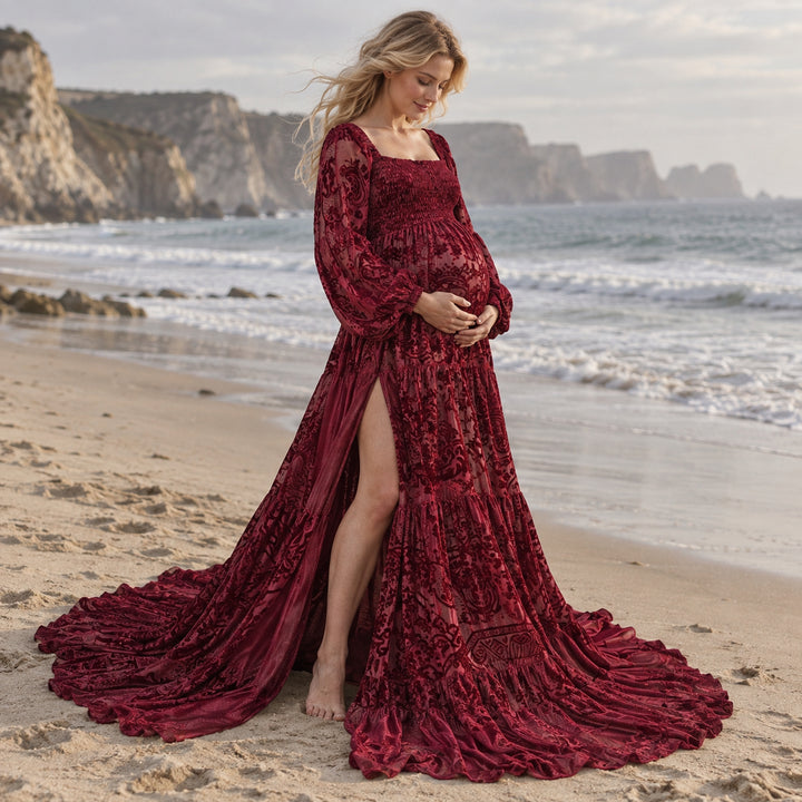 A pregnant woman in a Calypso red wine long-sleeve slit dress stands barefoot on a beach, hands on her belly, with sea and cliffs behind.
