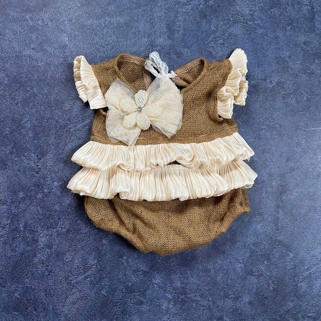 The flat lay shows a camel knit romper with double layer ruffles, a mesh flower, and lace ties on a dark blue background.