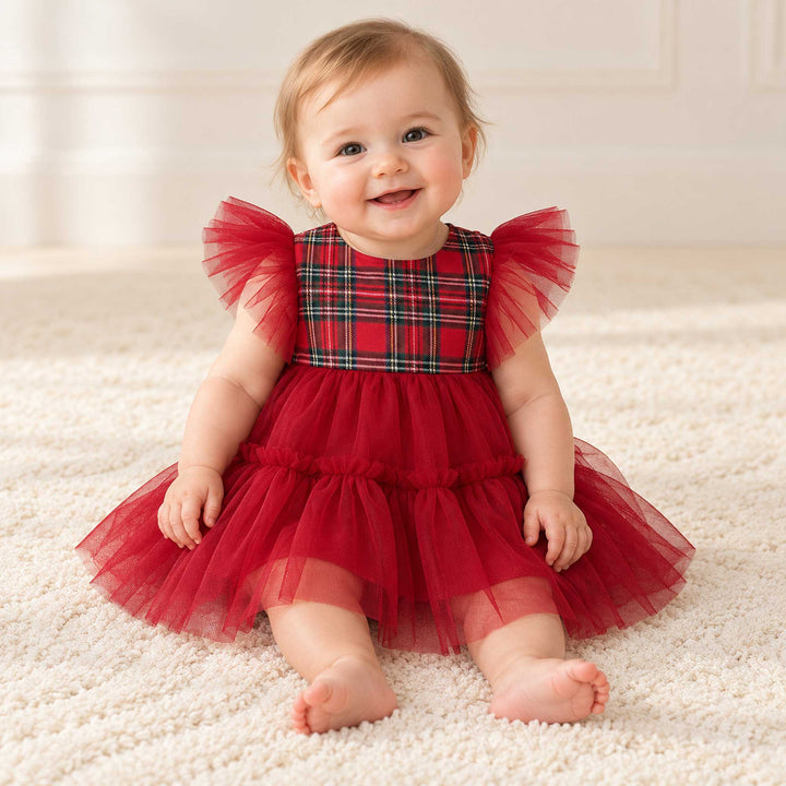 A baby in the Cerridwen dress sits on a light carpet facing the camera, with a white interior wall behind.