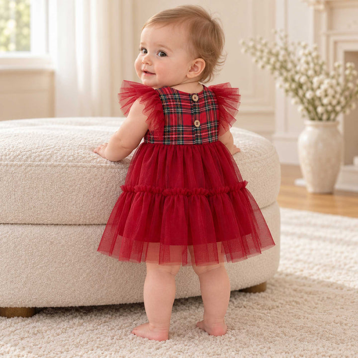 A baby in the Cerridwen dress stands with her back turned and head turned, one hand on an ottoman, buttons on the back, a vase on the right.