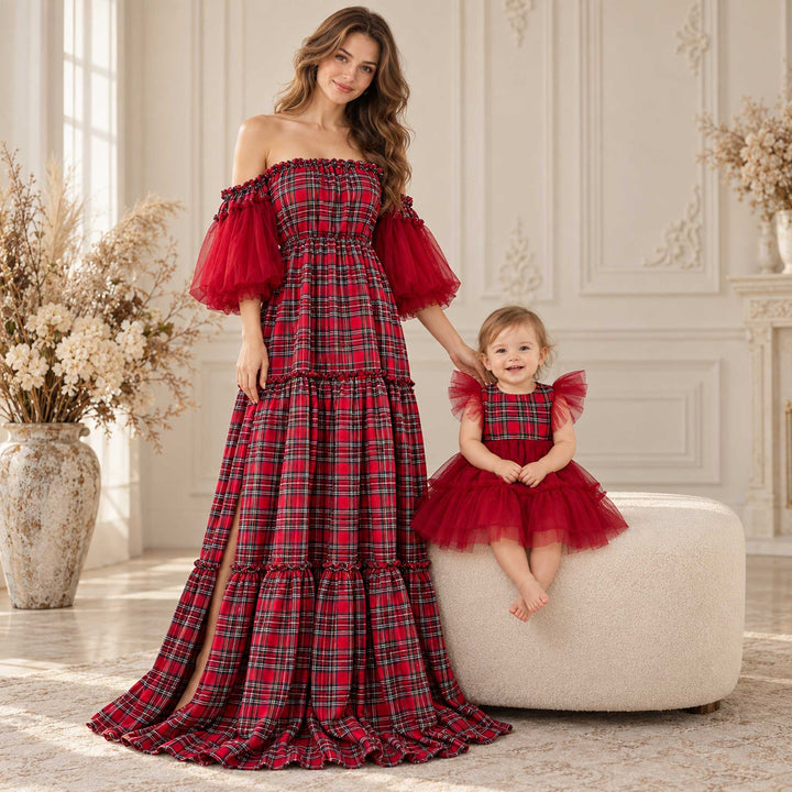 A woman in a charybdis plaid off-shoulder dress with red tulle sleeves rests a hand on a baby seated on an ottoman in a matching plaid tulle dress, indoors with vases and a paneled wall.