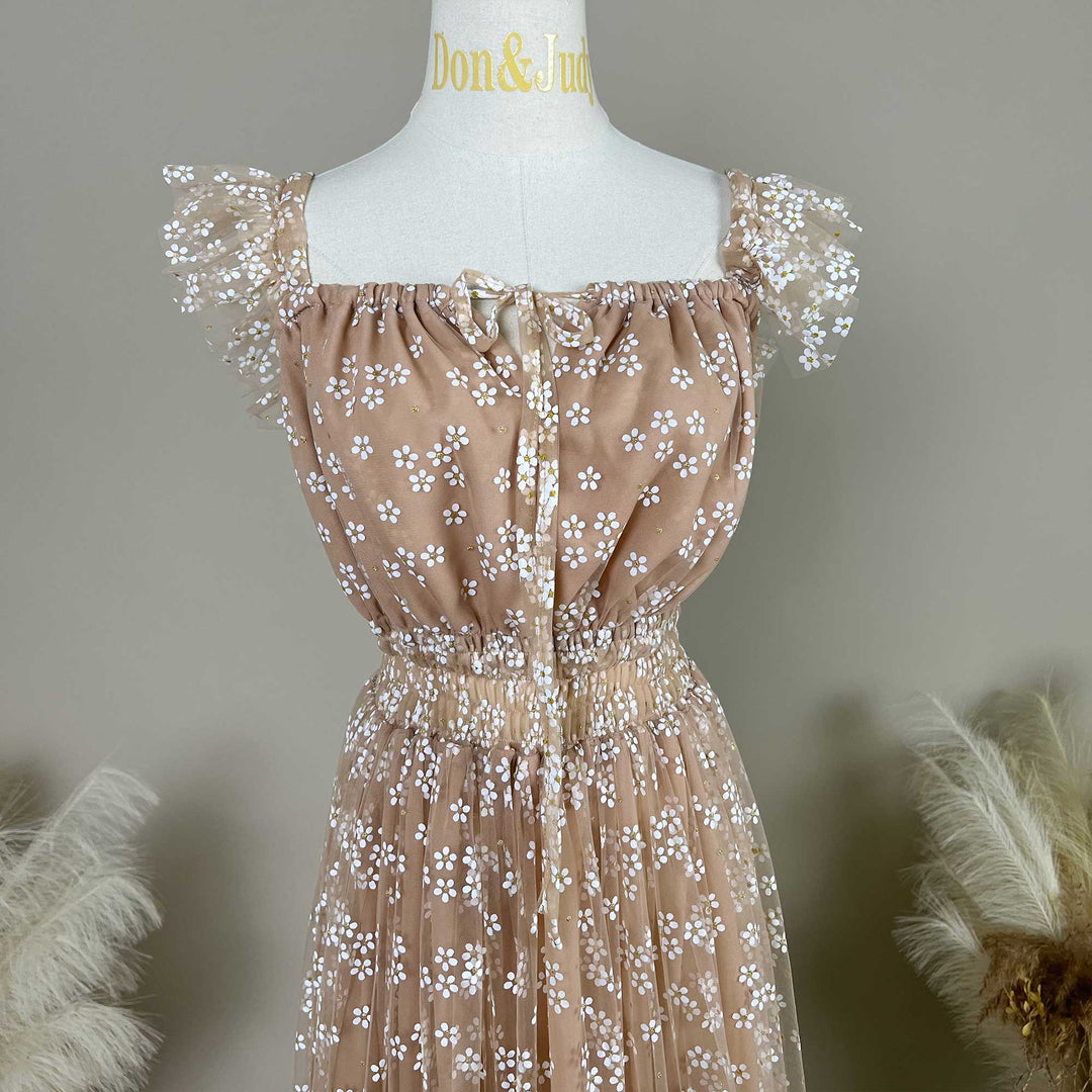 A close-up view of a khaki tulle maternity dress on a mannequin , featuring a square neckline with a delicate front tie, gathered elastic waist, and sheer ruffled cap sleeves adorned with white floral embroidery.