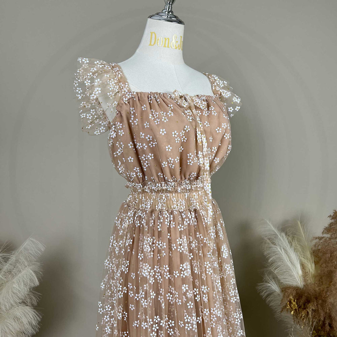 A close-up view of a khaki tulle maternity dress on a mannequin , featuring a square neckline with a delicate front tie, gathered elastic waist, and sheer ruffled cap sleeves adorned with white floral embroidery.
