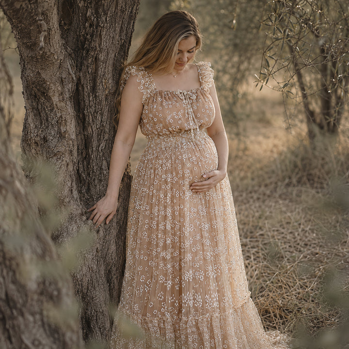 A pregnant woman leans against a tree trunk in a sunlit grove, wearing a khaki floral embroidered tulle maternity gown with ruffled square neckline and tiered skirt.