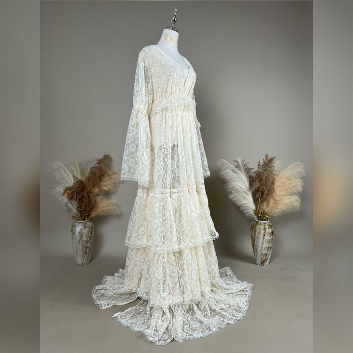 A Clito beige lace long-sleeve dress on a mannequin from an oblique angle, tiered skirt, train on the floor, a dried-flower vase on each side.