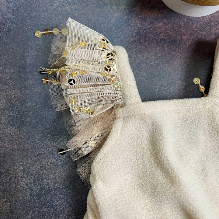 a close-up of a cream fleece baby suspender romper, showing one tulle ruffle strap and gold sequin strands, laid on a dark surface.