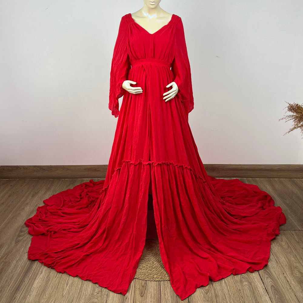 A Daphne red long-sleeved V-neck gown is displayed on a mannequin, with hands positioned on the simulated belly. The dress features a high front slit and its hem and train are spread out on the wooden floor.