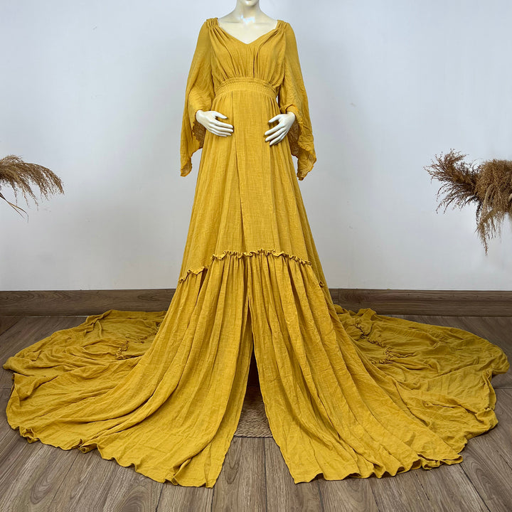 A Daphne yellow long-sleeved V-neck gown is displayed on a mannequin, with hands positioned on the simulated belly. The dress features a high front slit and its hem and train are spread out on the wooden floor.


