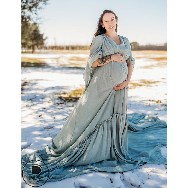 A pregnant woman in a Daphne light blue long-sleeve slit dress stands on snowy grass, hands on her belly, watermark at bottom left.