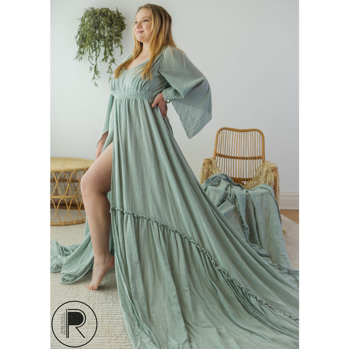 A woman in a sage long-sleeve slit dress stands barefoot on an indoor rug, one hand on her hip.