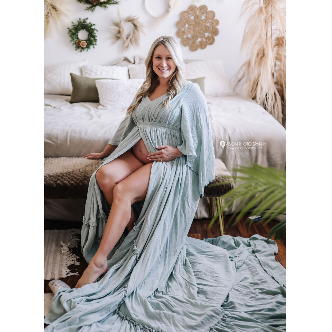 A woman sits on a bedroom bench wearing a Daphne ppale smokey blue gown with the center open, revealing her belly. Her hands are on her belly, and the hem lies on the floor.