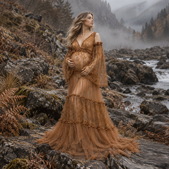 A woman stands on rocky riverbanks wearing a flowing lace maternity gown with dramatic off-the-shoulder sleeves and a full layered tulle skirt. 
