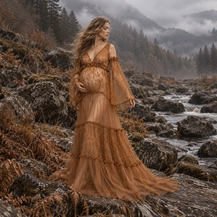 A pregnant woman stands on a rocky riverbank wearing a sheer, tiered rust tulle gown with long draped sleeves, set against a misty mountain forest and rushing stream background.