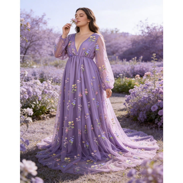 A woman smells a flower in a field, wearing a Dike purple tulle long-sleeve gown with floral embroidery and a train.