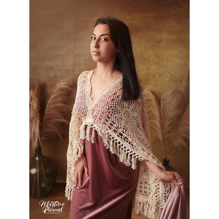 A woman wears Edwina beige crochet poncho over a KO-4 V-neck slip dress. Two bunches of dried flowers are placed in the background behind her。