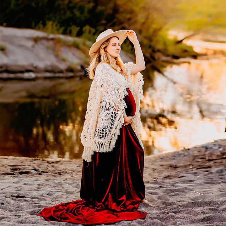 A pregnant woman wearing Edwina KO-14 velvet gown, a white crochet poncho, and a hat stands on a sandy ground near water. She holds her belly with one hand and touches her hat with the other.